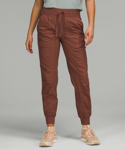 A person wearing jogger-style athletic pants in a muted terracotta brown color, paired with a light gray top and beige sneakers, against a plain light gray studio background.
