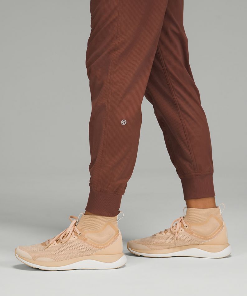 Close-up of a person's legs wearing brown jogger pants and beige athletic sneakers against a neutral gray studio background.