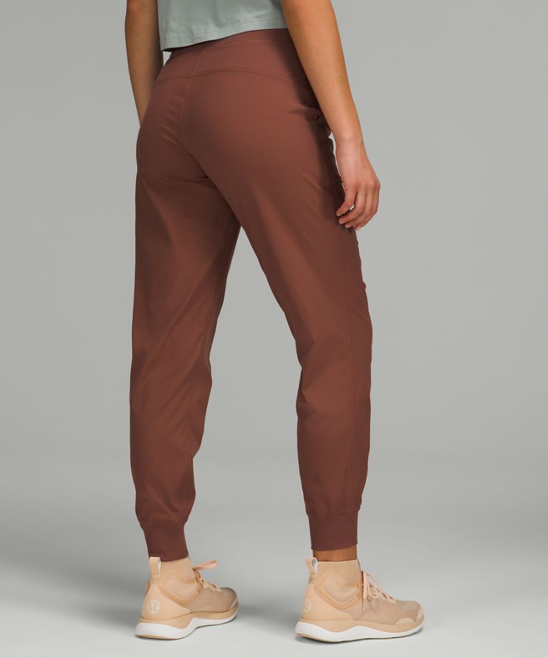 Rust brown jogger-style pants shown from the back on a model, paired with beige sneakers against a solid gray studio background.