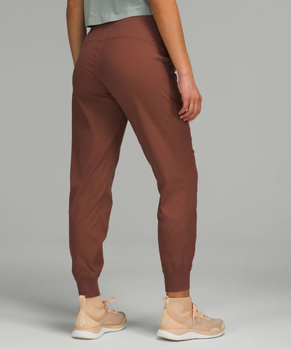 Rust brown jogger-style pants shown from the back on a model, paired with beige sneakers against a solid gray studio background.