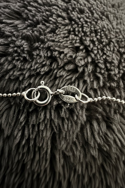 Close-up of a silver-toned chain bracelet with a lobster clasp and oval link detail, resting on a dark gray shaggy textured surface. The oval link is engraved with 'ITALY 925'.