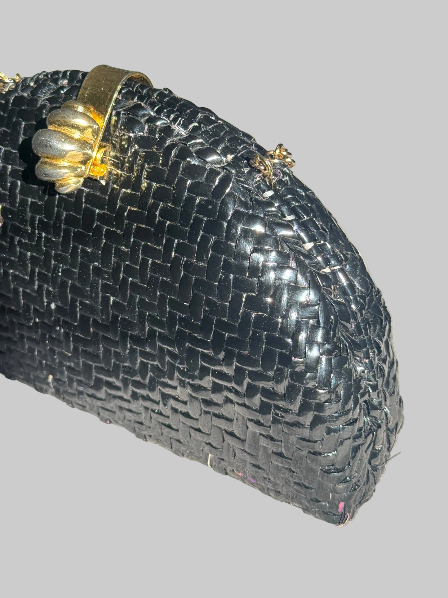 A close-up of a black woven clutch purse with a gold-tone metal clasp handle, resting on a textured light gray fabric surface.