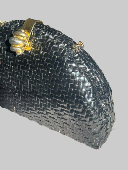 A close-up of a black woven clutch purse with a gold-tone metal clasp handle, resting on a textured light gray fabric surface.