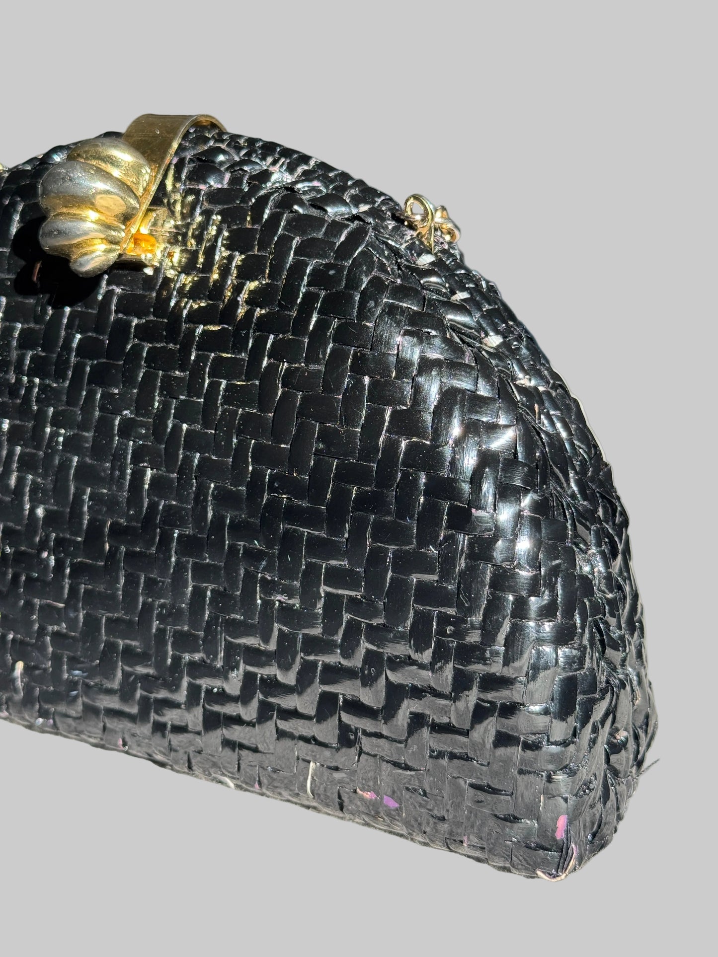 A close-up of a black woven clutch purse with a gold-tone decorative clasp on a light gray fabric surface.