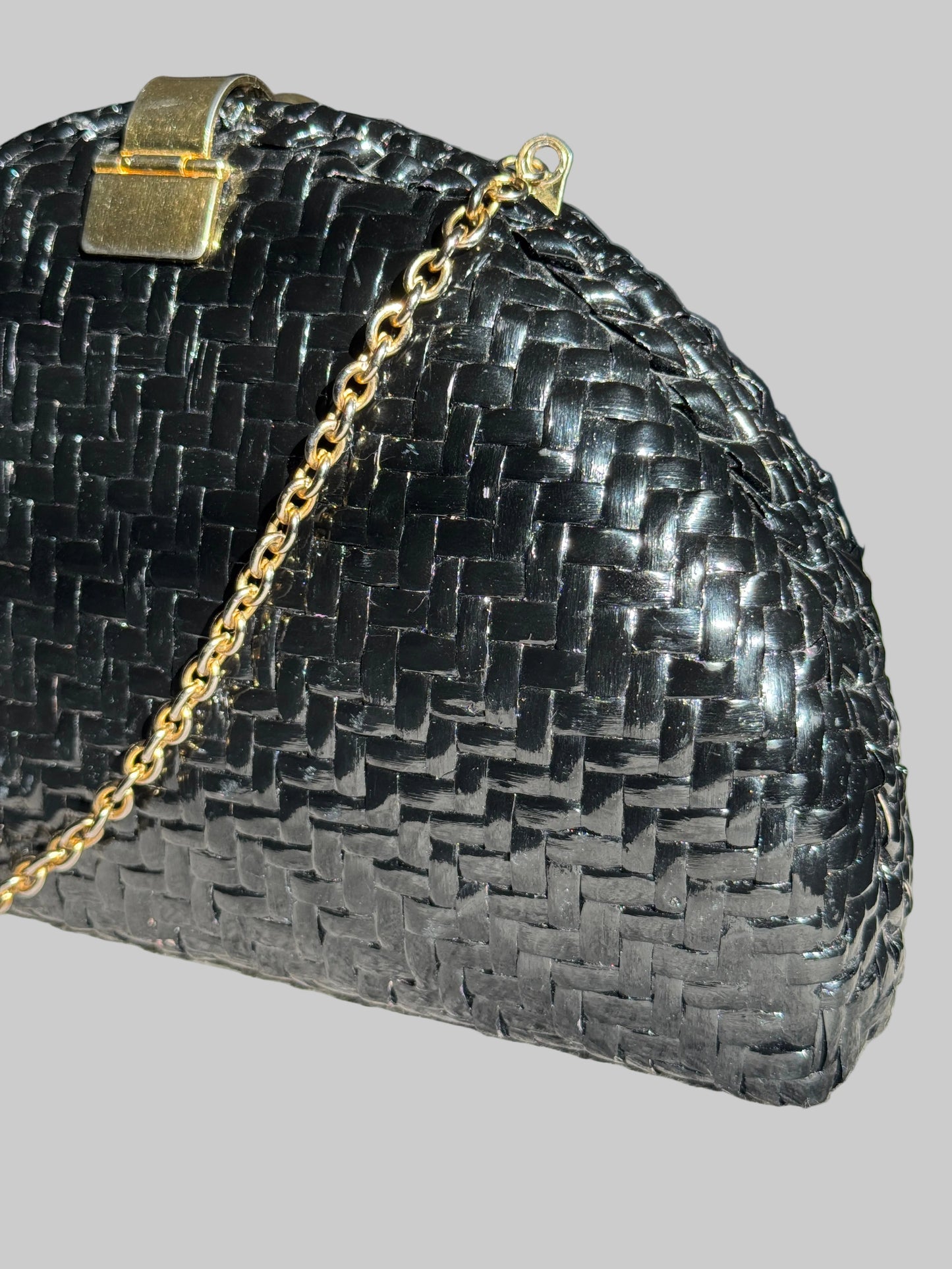 A close-up of a black woven leather clutch purse with a gold-tone chain strap and clasp, resting on a textured light gray surface.