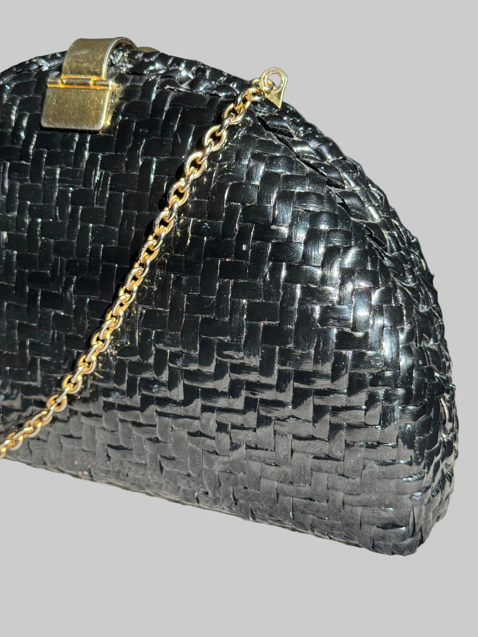 A close-up of a black woven leather clutch purse with a gold-tone chain strap and clasp, resting on a textured light gray surface.