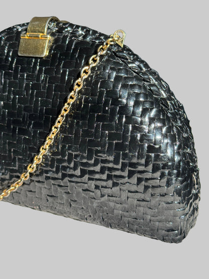 A close-up of a black woven leather clutch purse with a gold-tone chain strap and clasp, resting on a textured light gray surface.
