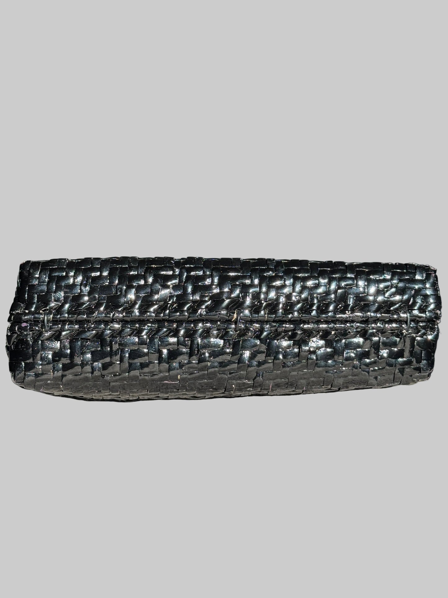 A black woven leather clutch bag with a textured, basket-like pattern, resting on a gray fabric surface with a blurred television in the background.
