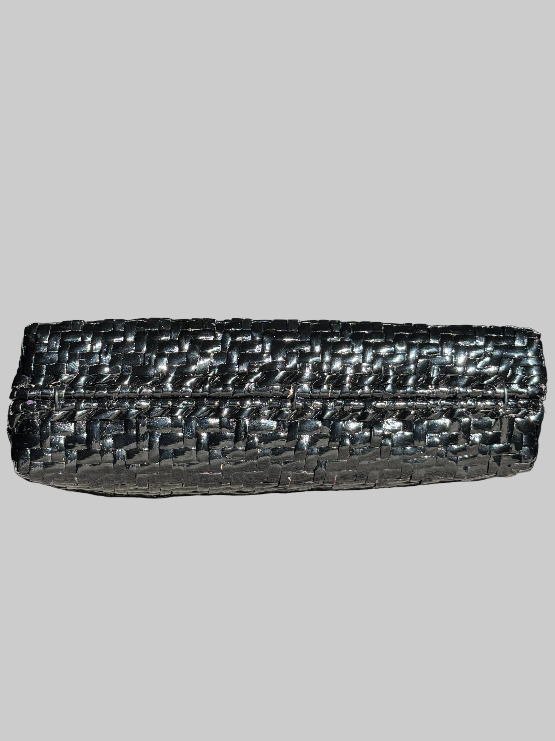 A black woven leather clutch bag with a textured, basket-like pattern, resting on a gray fabric surface with a blurred television in the background.