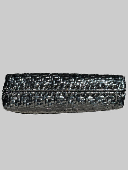 A black woven leather clutch bag with a textured, basket-like pattern, resting on a gray fabric surface with a blurred television in the background.