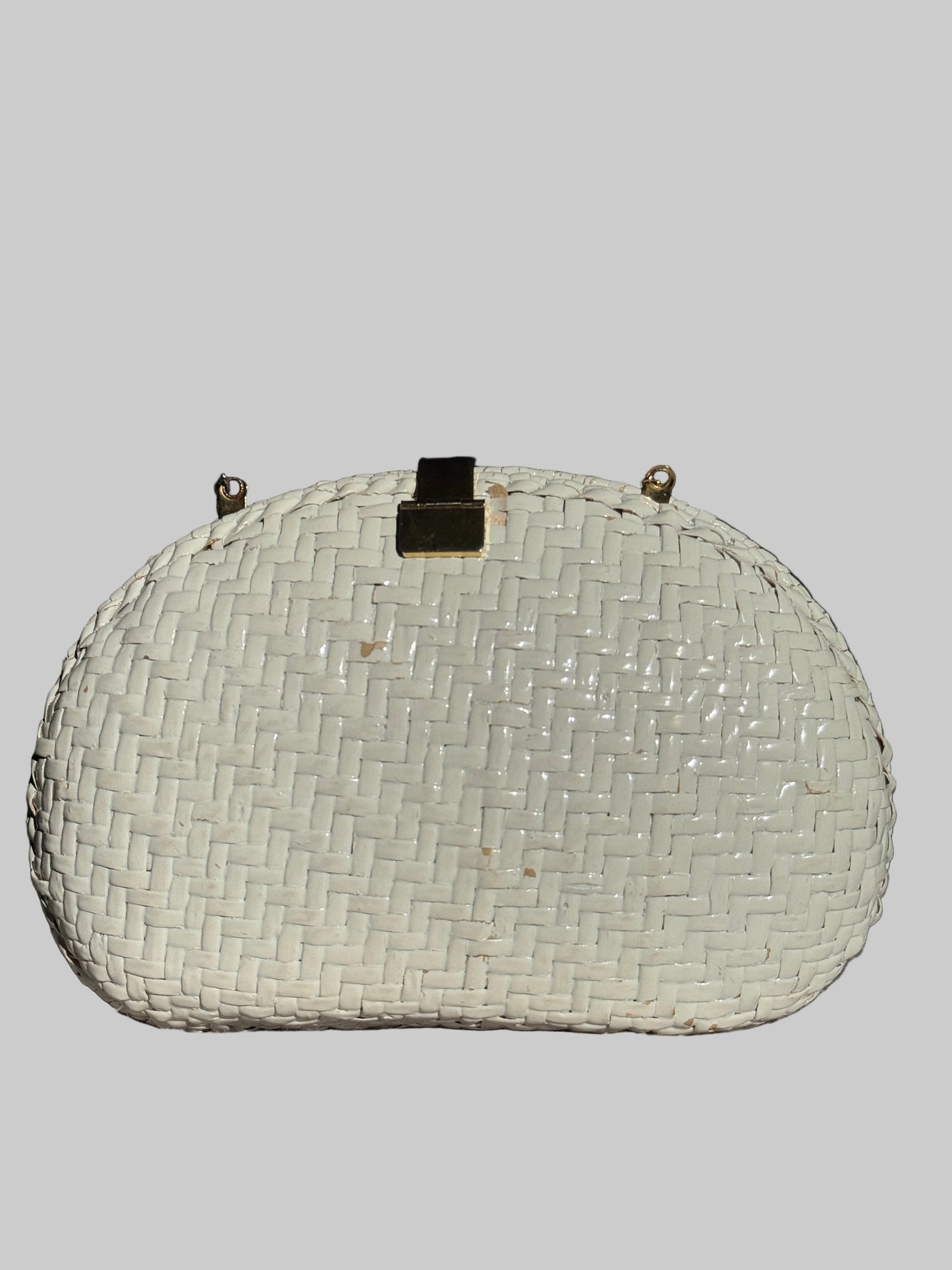 A white woven straw clutch purse with gold-tone hardware, resting on a textured gray surface against a dark, dimly lit background.