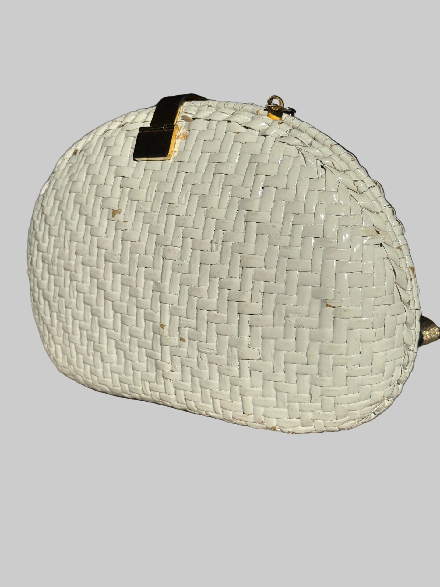 A white woven oval clutch purse with gold-tone hardware, resting on a textured gray surface.