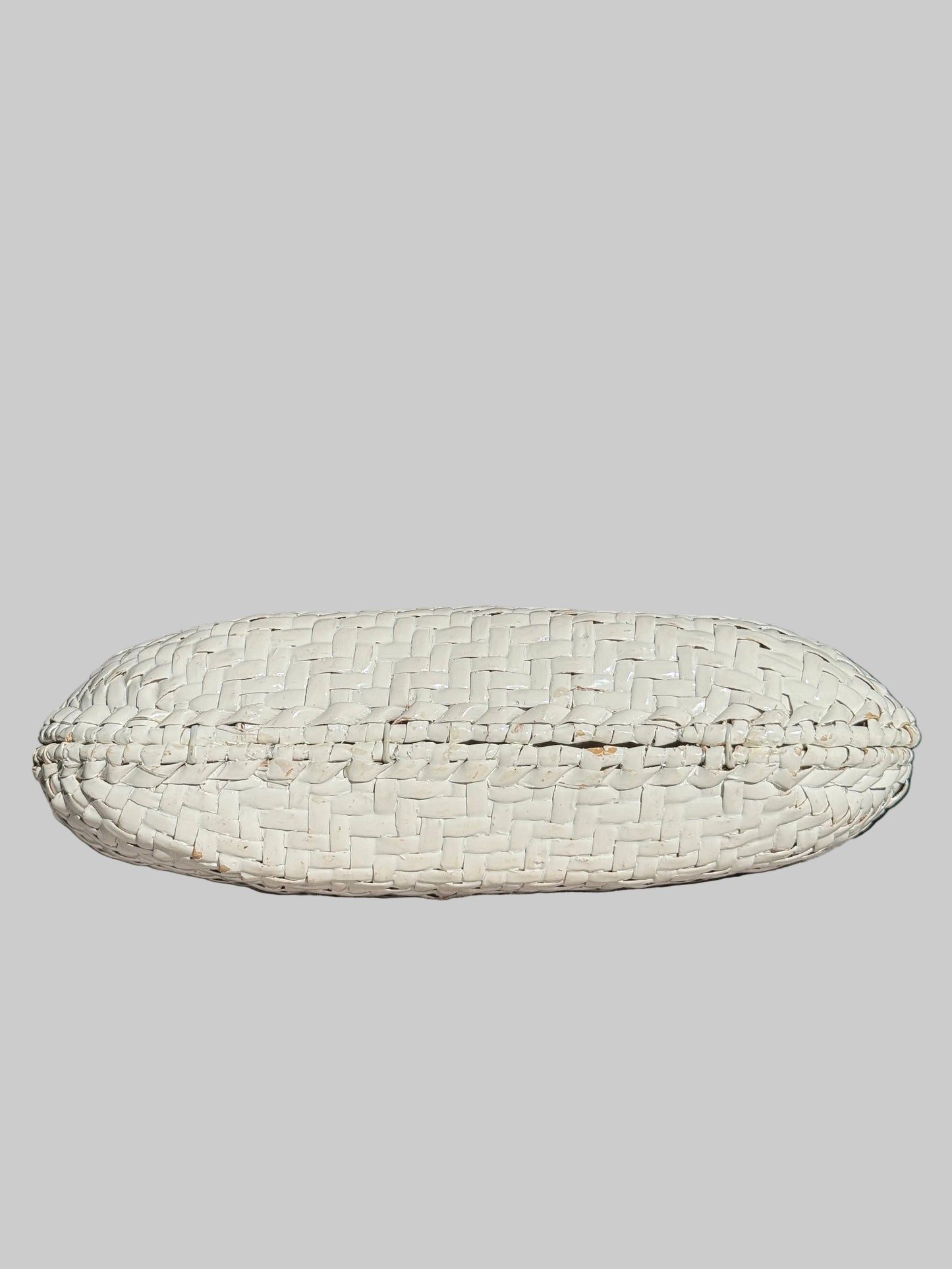 A white woven basket-style clutch or pouch with a textured, basketweave pattern, resting on a light gray fabric-covered surface with a dark, blurred background.