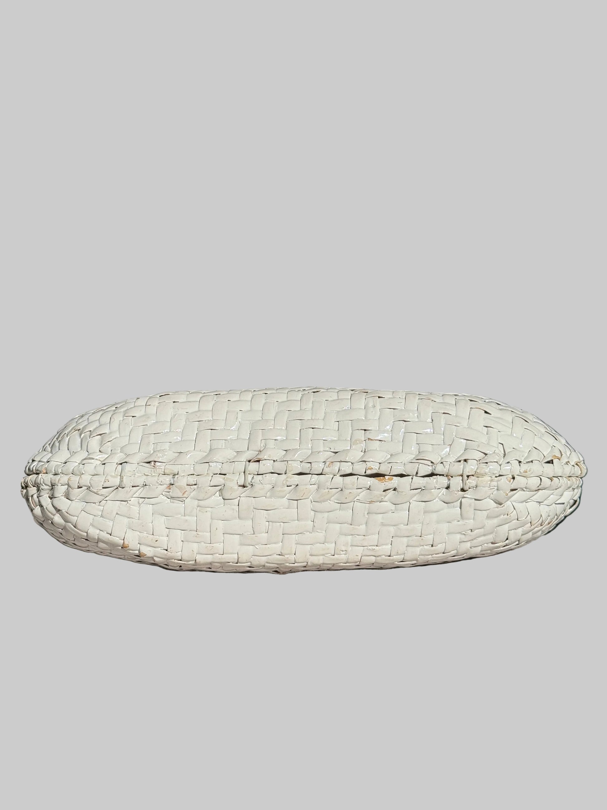 A white woven basket-style clutch or pouch with a textured, basketweave pattern, resting on a light gray fabric-covered surface with a dark, blurred background.