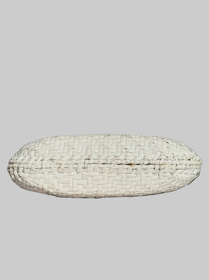 A white woven basket-style clutch or pouch with a textured, basketweave pattern, resting on a light gray fabric-covered surface with a dark, blurred background.