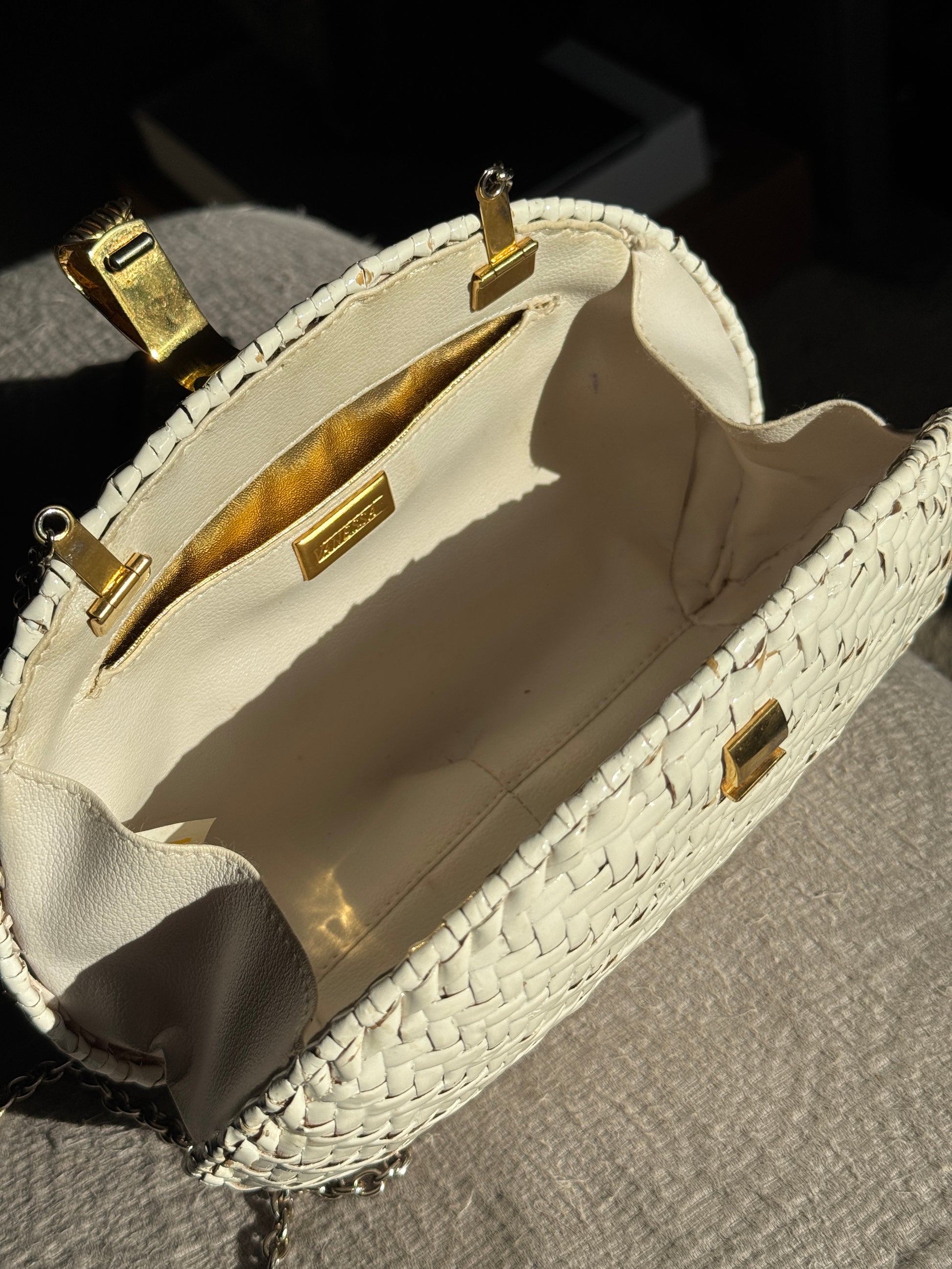 An off-white woven clutch handbag with gold-tone hardware, shown open to display its cream-colored interior lining and interior pocket.