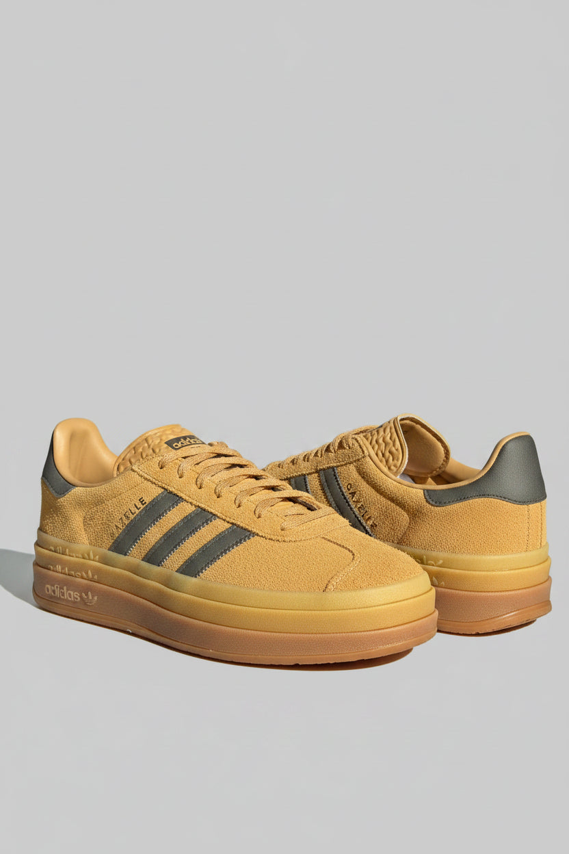 A pair of Adidas Gazelle platform sneakers in a yellow and brown colorway, displayed on a carpeted floor in front of a television and wall certificates.