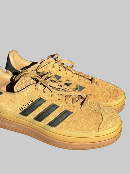 A pair of Adidas Gazelle sneakers in a warm tan suede and gum rubber colorway, featuring black three-stripes and 'GAZELLE' branding on the side, resting on a textured gray surface.