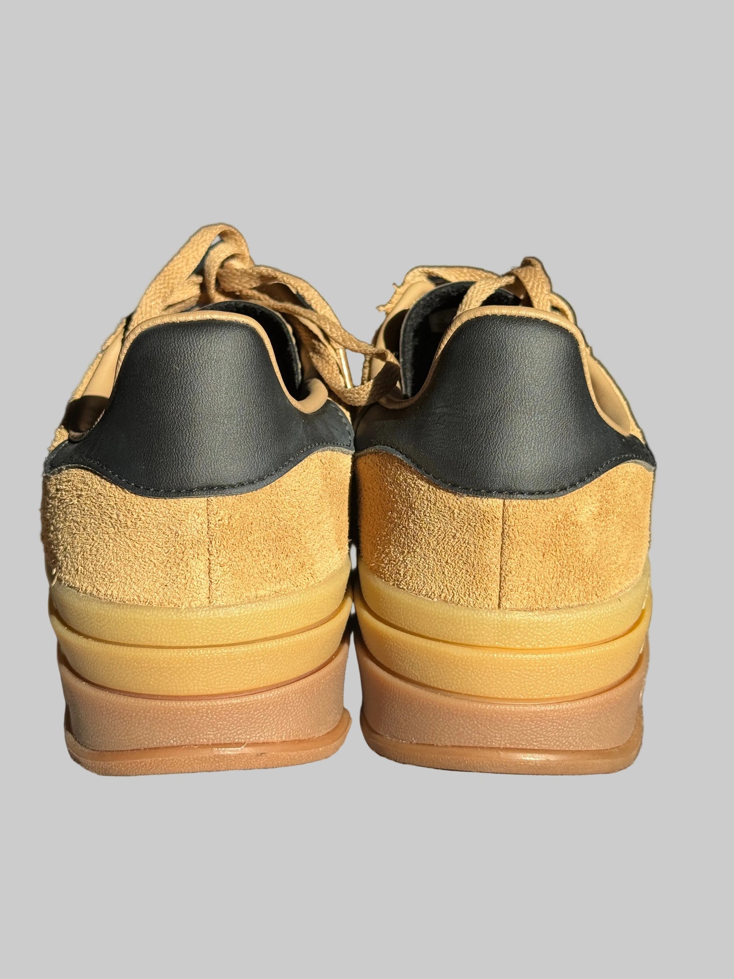 A pair of low-top sneakers shown from the back, featuring tan suede and black leather panels with a thick yellow and brown stacked sole, resting on a textured gray surface.