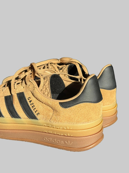 A pair of Adidas Gazelle sneakers in a tan suede and leather combination, featuring black three-stripes and a gum sole, resting on a textured gray surface.