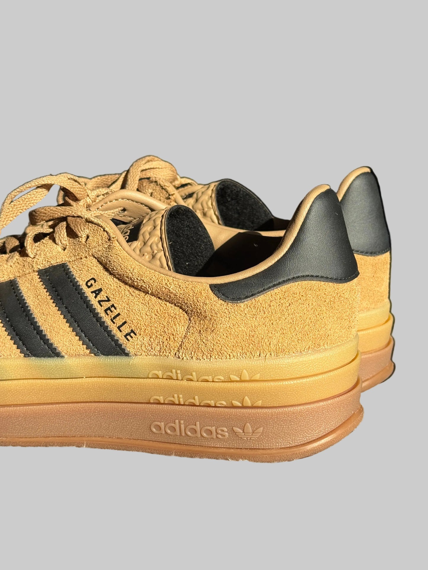A pair of Adidas Gazelle sneakers in a camel yellow suede and gum rubber colorway, featuring black leather stripes and heel tabs, resting on a textured gray surface.