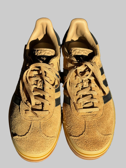 A pair of tan suede low-top sneakers with black side stripes and matching laces, photographed from above on a gray textured surface with direct sunlight and shadows.