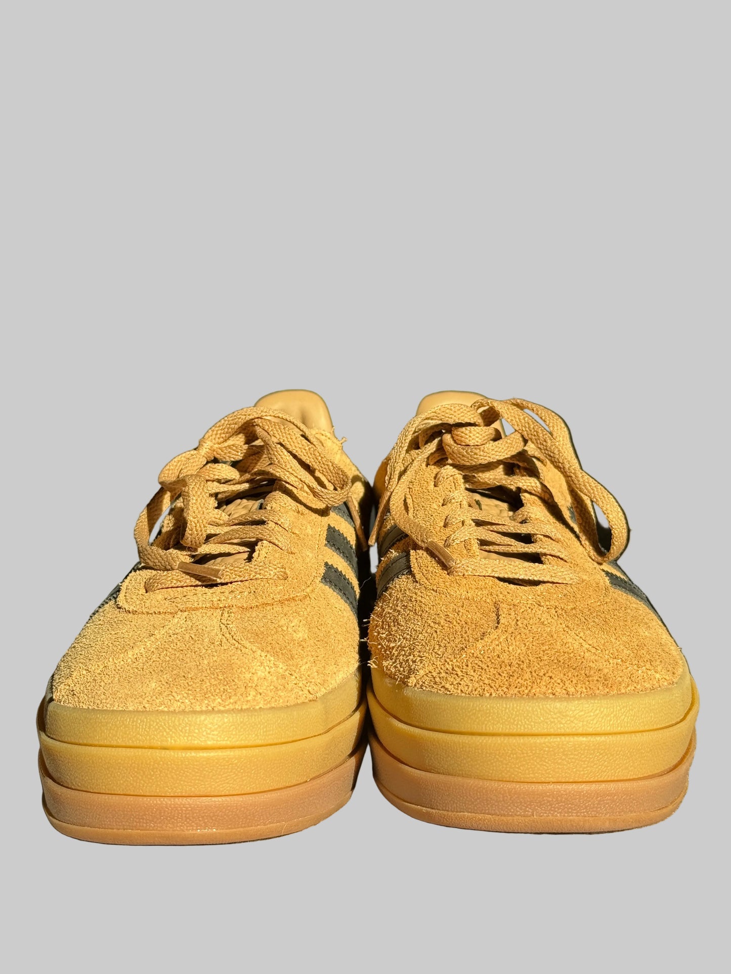 A pair of low-top platform sneakers in mustard yellow suede with dark green stripes and thick yellow-and-brown gum soles, resting on a textured surface with a television in the background.