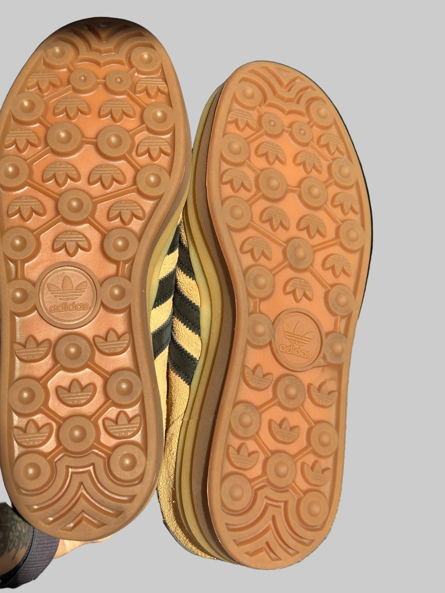 Close-up of the soles of two Adidas Trefoil sneakers, showing the gum rubber tread pattern and the embossed Adidas Trefoil logo, with a yellow and black striped side panel visible between the shoes.