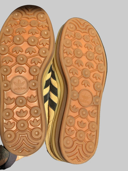 Close-up of the soles of two Adidas Trefoil sneakers, showing the gum rubber tread pattern and the embossed Adidas Trefoil logo, with a yellow and black striped side panel visible between the shoes.