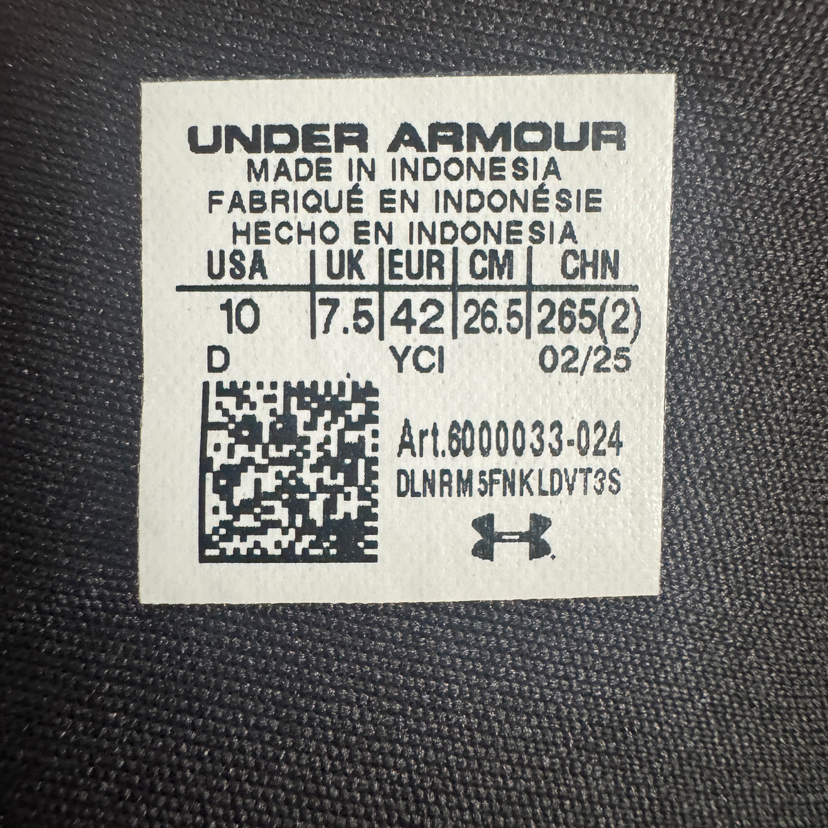 A close-up of a white Under Armour size label sewn onto black textured fabric, showing size information and a small Under Armour logo.