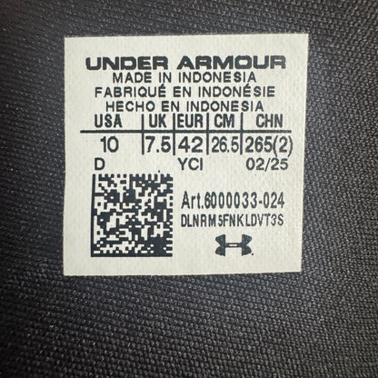 A close-up of a white Under Armour size label sewn onto black textured fabric, showing size information and a small Under Armour logo.