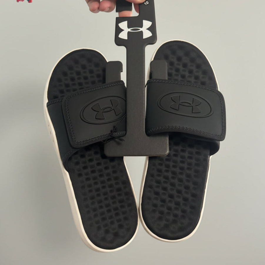 A pair of black Under Armour slide sandals held by a black retail hanger in a person's hand, with a home interior visible in the background.