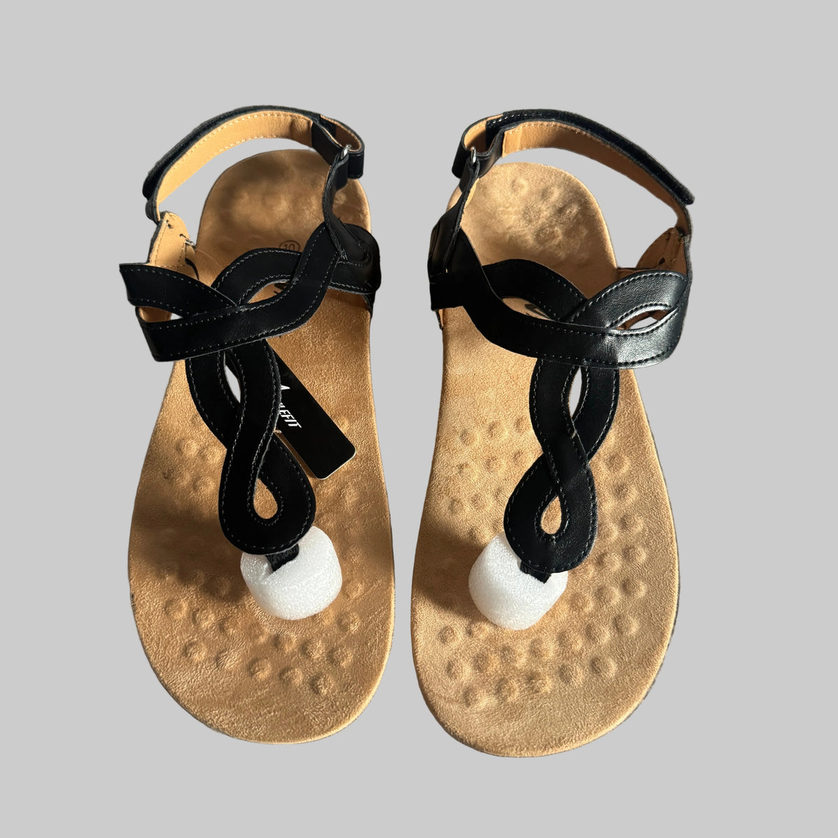 A pair of black leather T-strap sandals with textured tan footbeds and white pom-pom accents, laid flat on a gray textured fabric background.