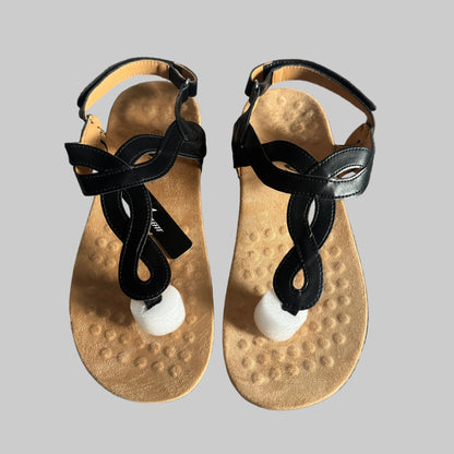 A pair of black leather T-strap sandals with textured tan footbeds and white pom-pom accents, laid flat on a gray textured fabric background.