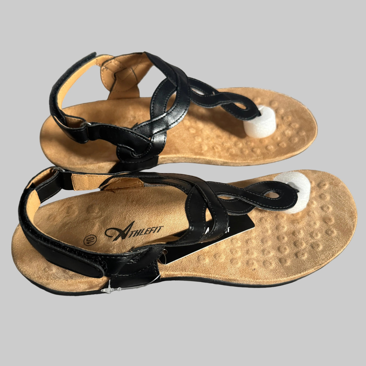 A pair of black leather strappy sandals with textured tan footbeds and white toe supports, displayed on a gray textured fabric background.