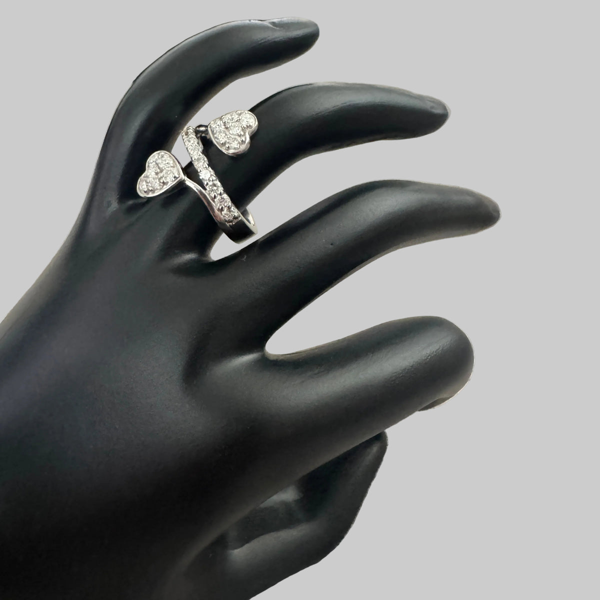 A silver-toned ring with two heart-shaped pavé settings, displayed on a black mannequin hand over a textured light gray fabric background.