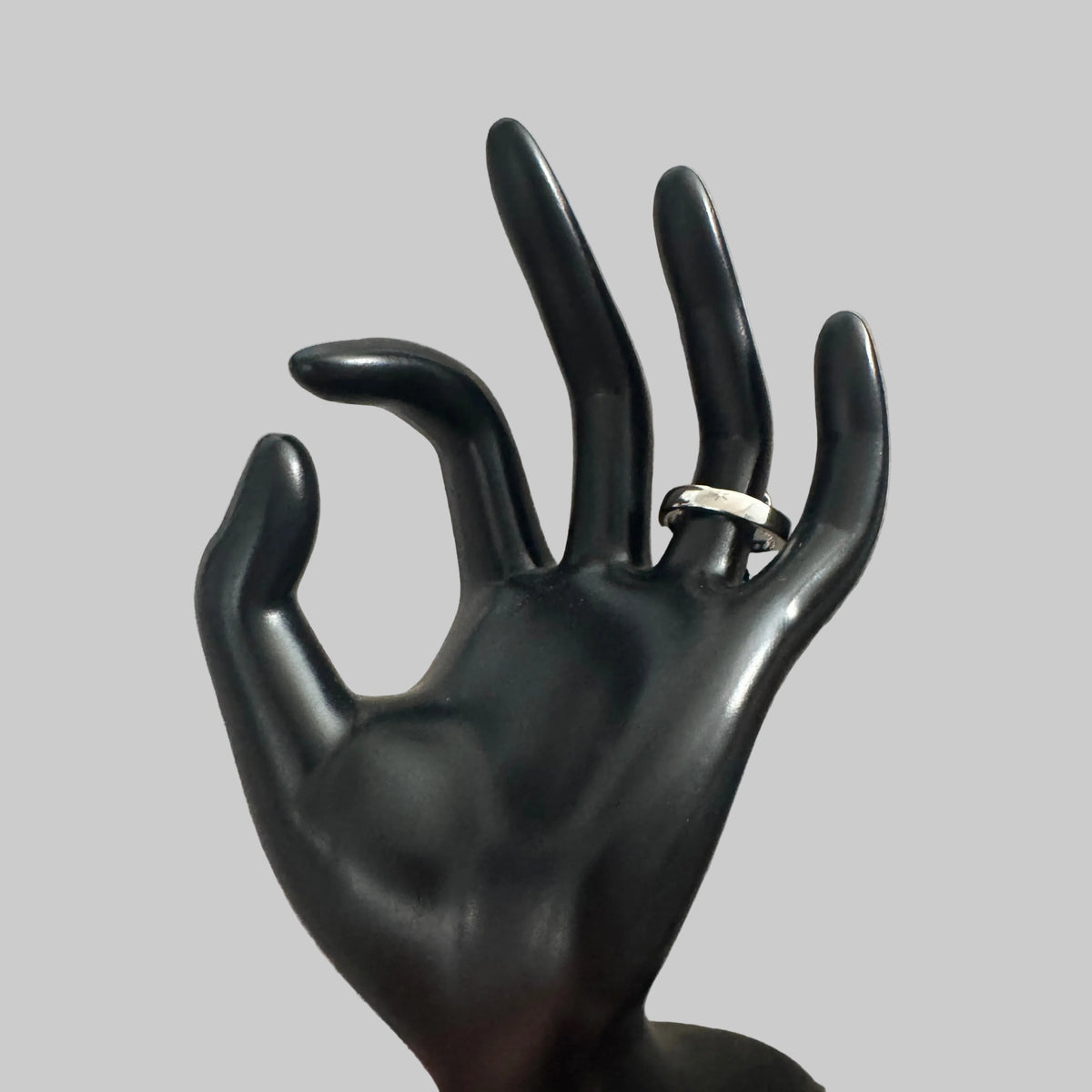 A silver-toned band ring displayed on a glossy black hand-shaped jewelry stand against a textured light gray fabric background.