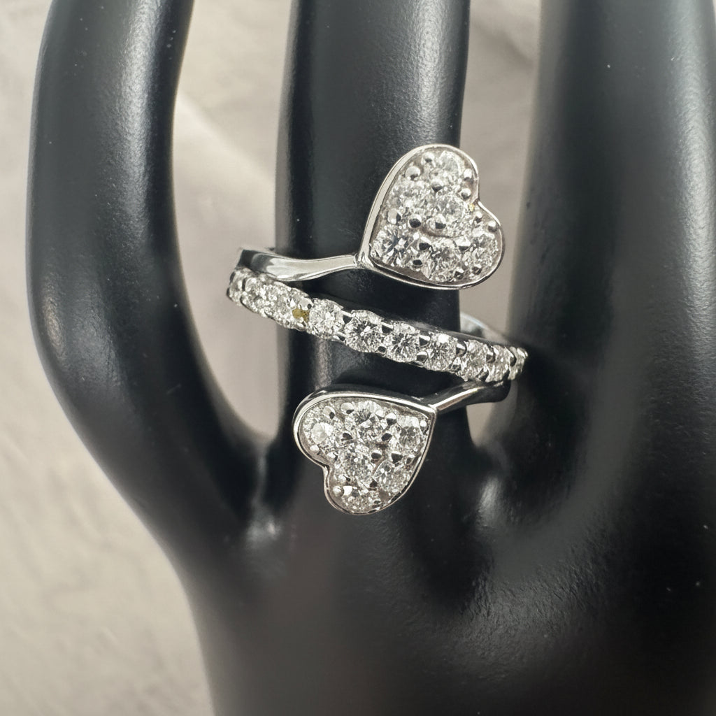 A silver-toned ring with two heart-shaped pavé settings and a matching pavé band, displayed on a black jewelry stand.