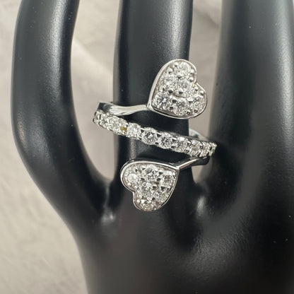 A silver-toned ring with two heart-shaped pavé settings and a matching pavé band, displayed on a black jewelry stand.