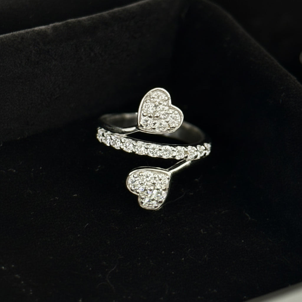 A silver-toned open ring featuring two heart-shaped pavé settings, displayed in a black velvet ring box.