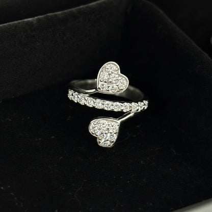 A silver-toned open ring featuring two heart-shaped pavé settings, displayed in a black velvet ring box.