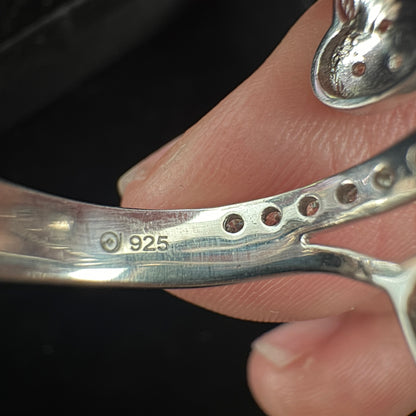 Close-up of a silver-toned metal bracelet held against skin, showing a sterling silver hallmark stamped '925' and small circular cutouts along the band.