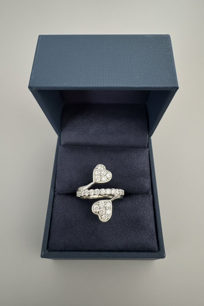 A silver ring with two heart-shaped pavé settings, displayed inside an open dark blue velvet jewelry box on a textured gray fabric background.