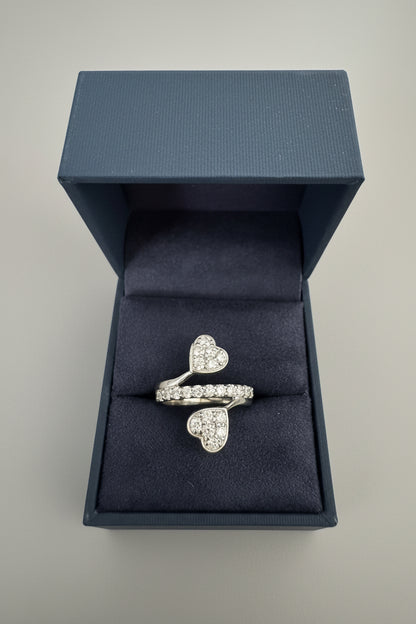 A silver ring with two heart-shaped pavé settings, displayed inside an open dark blue velvet jewelry box on a textured gray fabric background.