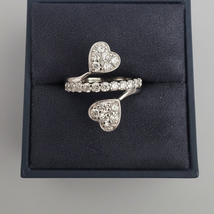 A silver-toned open-shelf ring box with two rings displayed inside. The front ring features a pavé band with a heart-shaped cluster of clear stones, and the rear ring has a pavé band with a separate dangling heart-shaped cluster.