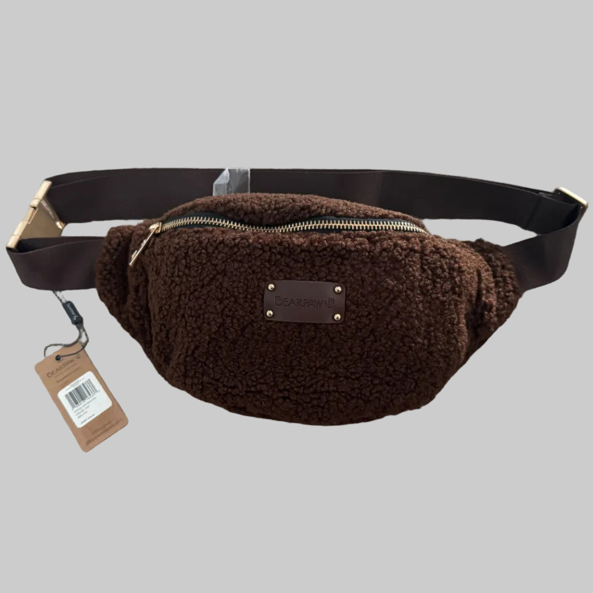 A brown sherpa fleece fanny pack with a matching adjustable strap and a small brown leather brand patch, shown on a gray carpet with its original paper tag attached.