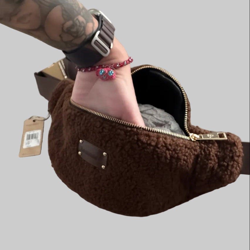 A brown fuzzy faux-fur crossbody bag with a dark brown strap and gold-tone zipper, shown on a beige carpet with a person's hand and tattooed arm resting inside the bag.