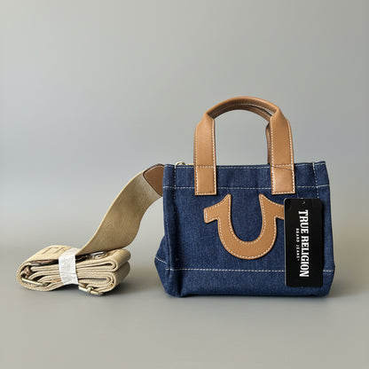 A blue denim tote bag with tan leather handles and a large tan logo emblem on the front, shown on a dark couch with a matching rolled strap beside it. A black hangtag reads True Religion.