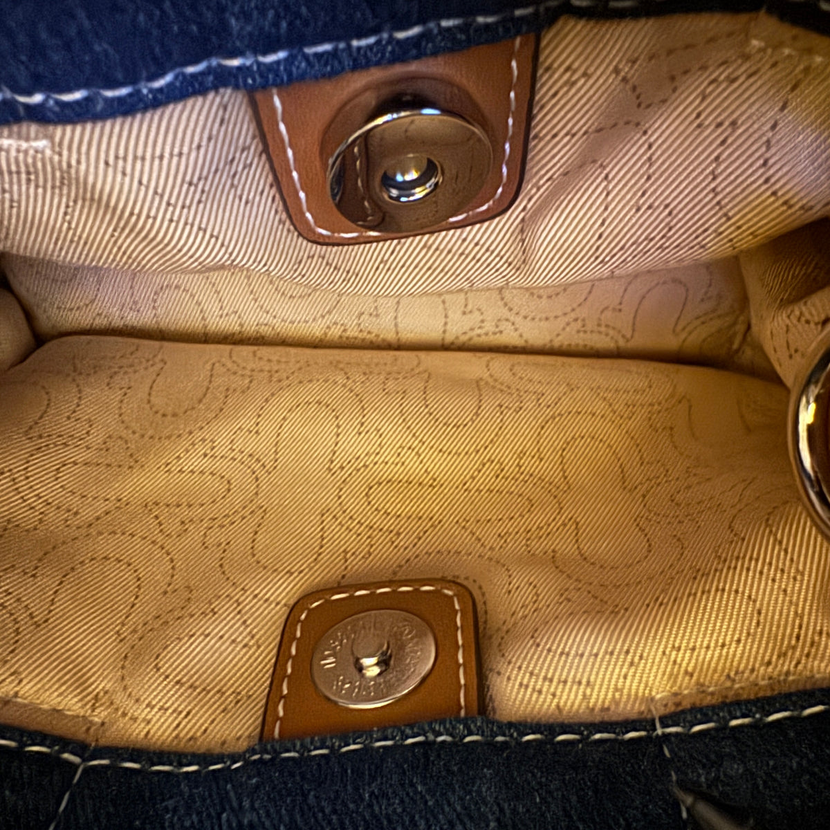 Close-up interior view of a handbag showing the tan patterned fabric lining, white contrast stitching, and brown leather snap closures with metal hardware.