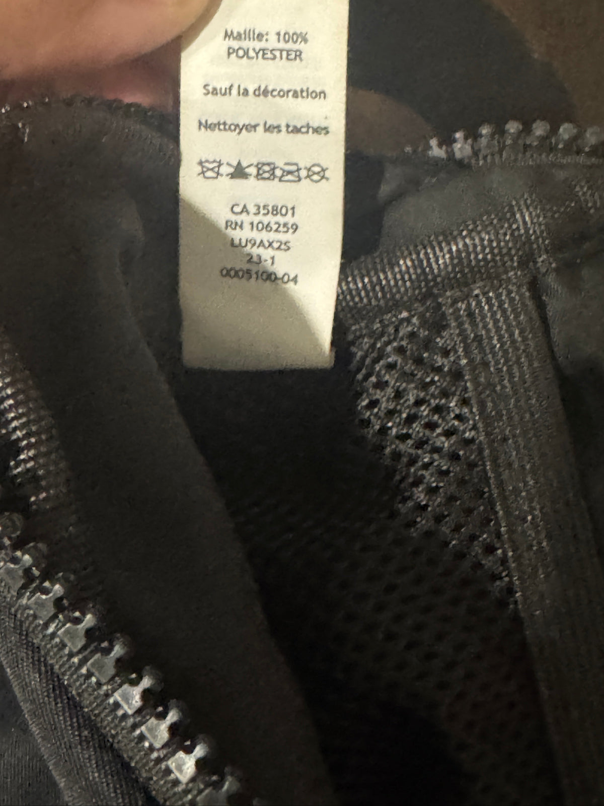 Close-up of a garment's interior label reading 'Molle: 100% POLYESTER' in French, attached to a black mesh zipper compartment.